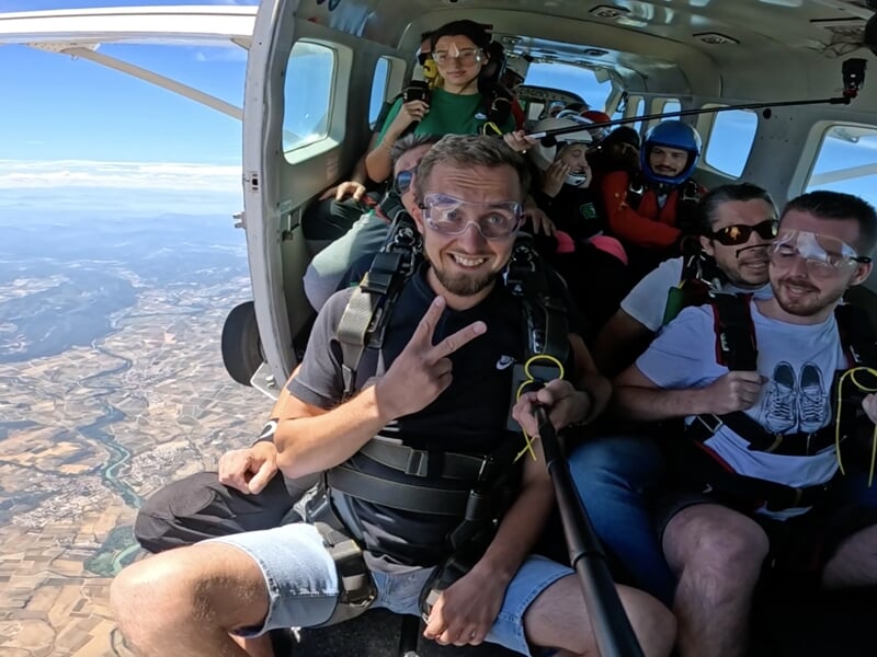 Groupe de parachutistes à bord d'un avion avant le saut, assis près de la porte ouverte avec une vue imprenable sur le paysage en contrebas. Un homme en premier plan fait un signe de victoire en souri