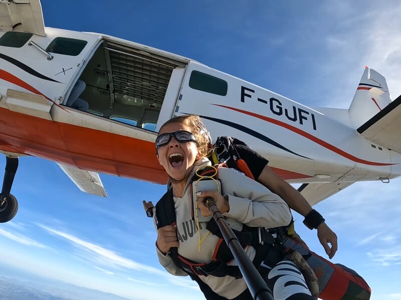 Sortie d’avion spectaculaire pour un saut en parachute en tandem. Une femme euphorique, équipée d’un harnais, crie de joie en quittant l’appareil avec son instructeur. L’avion blanc et rouge est encor