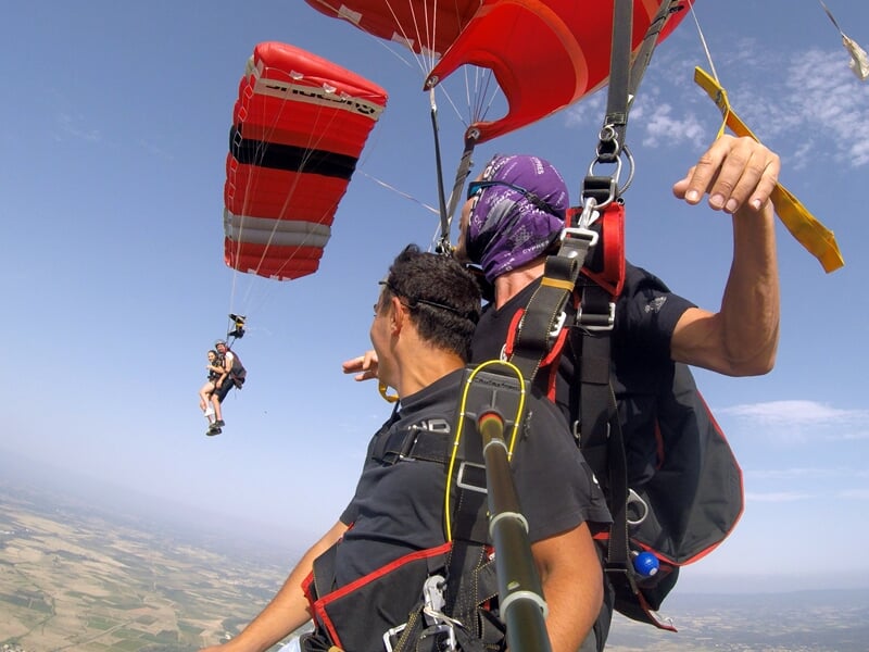 Descente sous parachute rouge et noir avec deux parachutistes en tandem observant un autre duo à proximité. Sensation de liberté et d’apesanteur dans le ciel avec une vue imprenable sur la campagne en