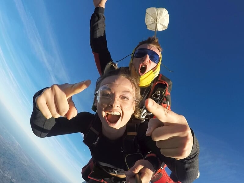Chute libre en tandem avec une femme radieuse pointant la caméra, accompagnée de son instructeur criant d’enthousiasme. Un petit parachute extracteur est visible en arrière-plan, flottant dans le ciel