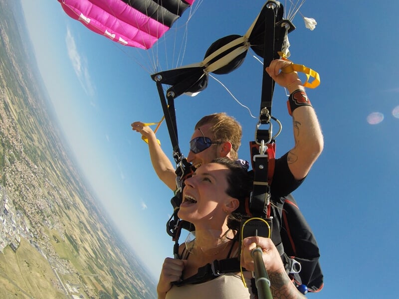 Phase de descente sous parachute biplace avec une voile rose et noire. La passagère, cheveux au vent, exprime sa joie tandis que son instructeur gère la trajectoire. Soleil éclatant et paysage urbain 