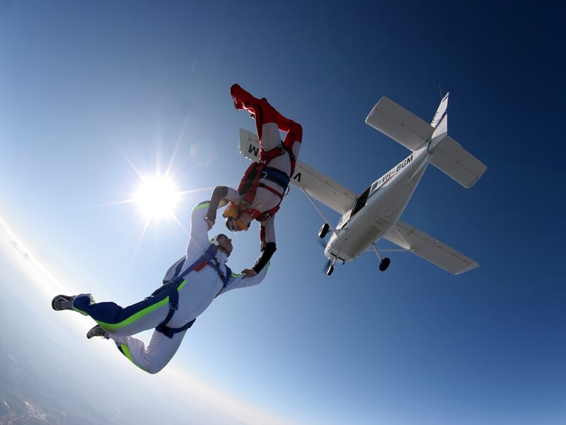 Sortie spectaculaire d’un avion en plein ciel, deux parachutistes en formation effectuent une figure synchronisée, se tenant par les mains. L’un est tête en bas avec une combinaison rouge et blanche,