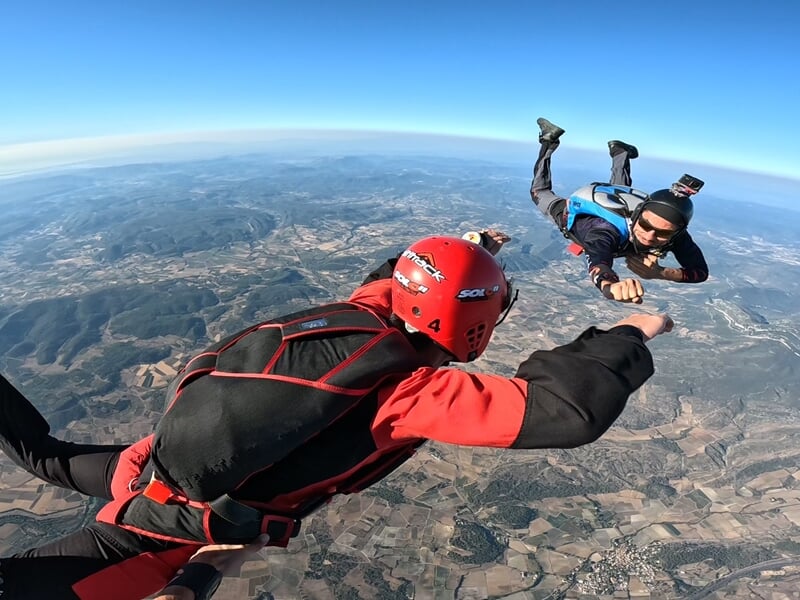 Rencontre aérienne entre deux parachutistes lors d’une chute libre. L’un tend le poing vers l’autre, équipé d’une caméra sur son casque, dans un geste de connexion et de maîtrise du vol en formation.