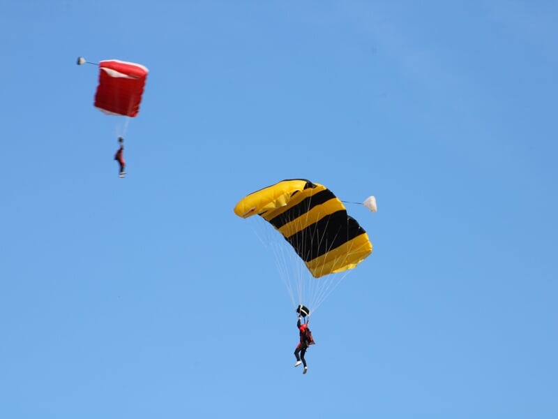 Descente sous parachute avec deux voiles distinctes : une rouge en arrière-plan et une jaune et noire en premier plan. Les parachutistes contrôlent leur atterrissage sous un ciel bleu dégagé.