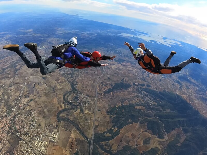 Trois parachutistes en pleine chute libre au-dessus d’un paysage vallonné avec une vue sur la mer. Deux d’entre eux sont en tandem, tandis qu’un troisième, vêtu d’une combinaison orange et noire, vole