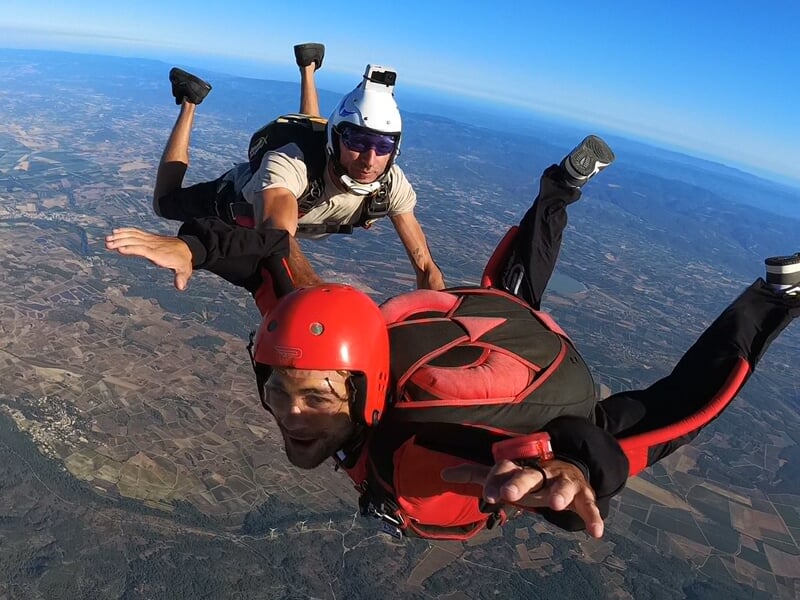 Chute libre en duo avec un parachutiste en combinaison rouge et noire, casque rouge, souriant face à la caméra, et un instructeur équipé d’un casque blanc et d’une caméra embarquée. Vol en formation a