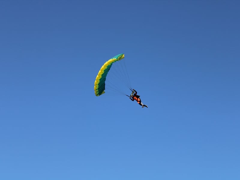 Parachutiste sous une voile jaune et verte descendant dans un ciel bleu dégagé. Position dynamique avec une légère inclinaison du corps, maîtrisant la trajectoire avant l’atterrissage.