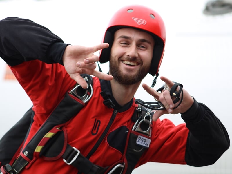 Portrait d’un parachutiste souriant en combinaison rouge et noire, casque rouge, effectuant un signe enthousiaste avec les mains. Joie et adrénaline après un saut réussi.