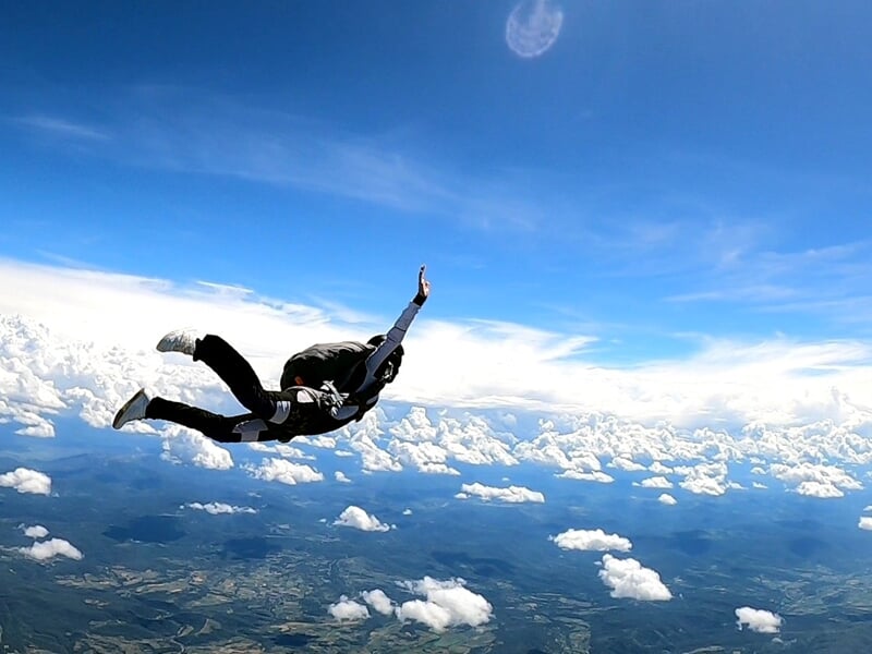 Parachutiste en pleine chute libre au-dessus d’un paysage montagneux et nuageux, bras levé vers le ciel. Sensation de liberté dans un ciel bleu éclatant parsemé de nuages.