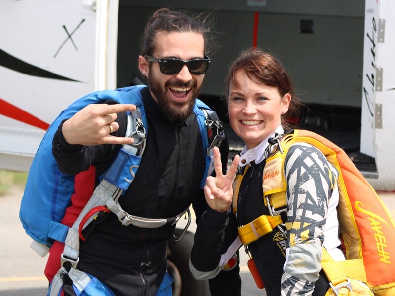 Deux parachutistes souriants devant un avion, portant leurs harnais et sacs de parachute. L’homme en combinaison noire et bleue fait un signe rock’n’roll, tandis que la femme en équipement jaune et no
