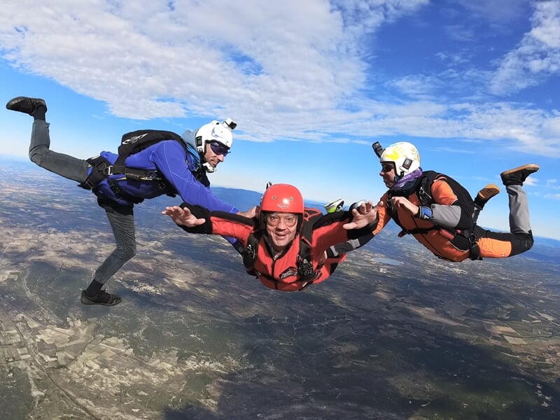 Vol en formation à trois parachutistes en pleine chute libre, avec une interaction dynamique. Le sauteur central en combinaison rouge et noire est entouré de deux autres en bleu et orange, tous maîtri