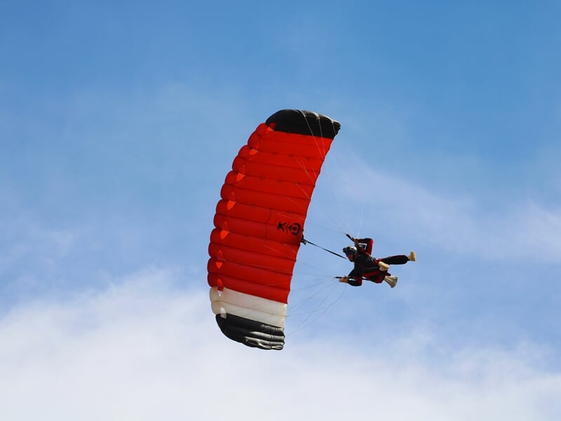 Parachutiste sous une voile rouge et noire en pleine descente, adoptant une posture dynamique. Ciel légèrement nuageux en arrière-plan, offrant un contraste avec le parachute vif.