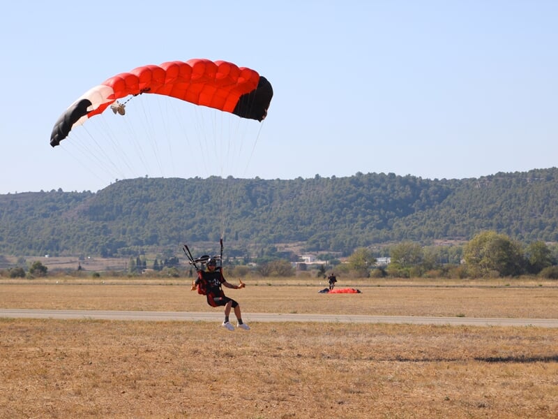 Atterrissage en douceur d’un parachutiste sous une voile rouge et noire sur un terrain sec, avec des collines verdoyantes en arrière-plan. Une autre personne ayant déjà atterri est visible au loin.