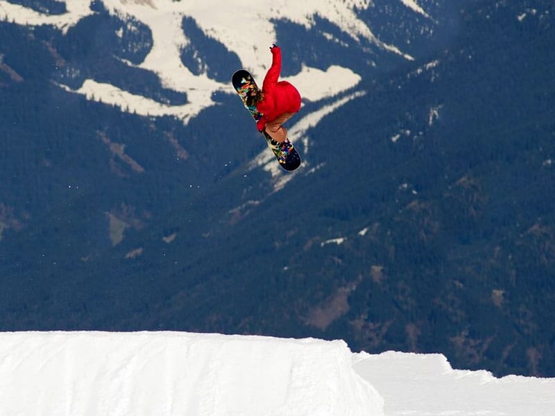 Un snowboardeur en combinaison rouge exécutant un saut acrobatique élevé avec des montagnes enneigées en arrière-plan
