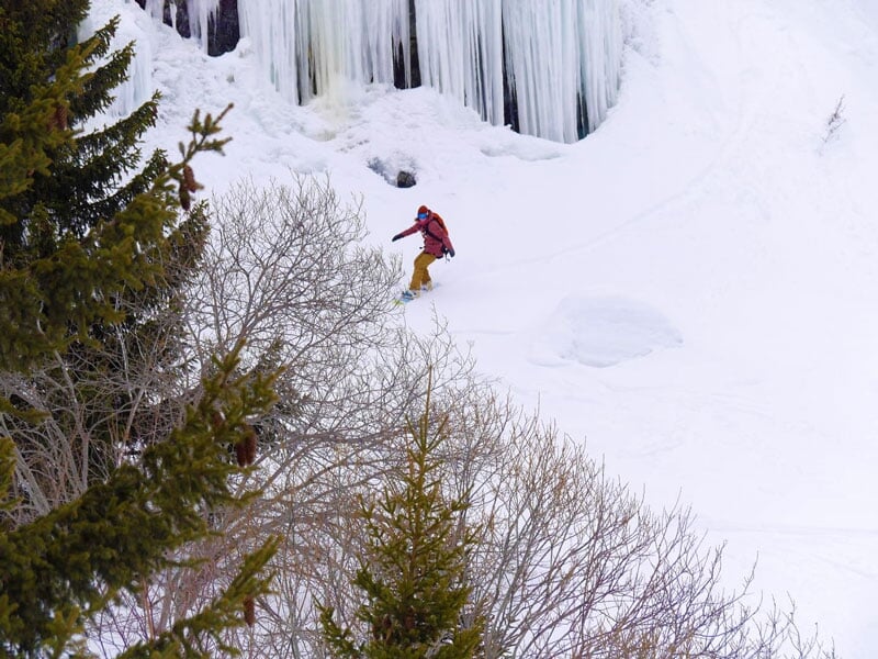Snowboardeur descendant une pente raide enneigée près d'une cascade glacée dans un environnement montagneux