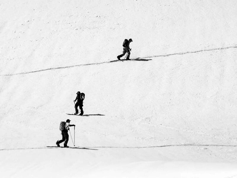 Trois randonneurs en snowboard gravissant une pente enneigée, dans un paysage hivernal en noir et blanc