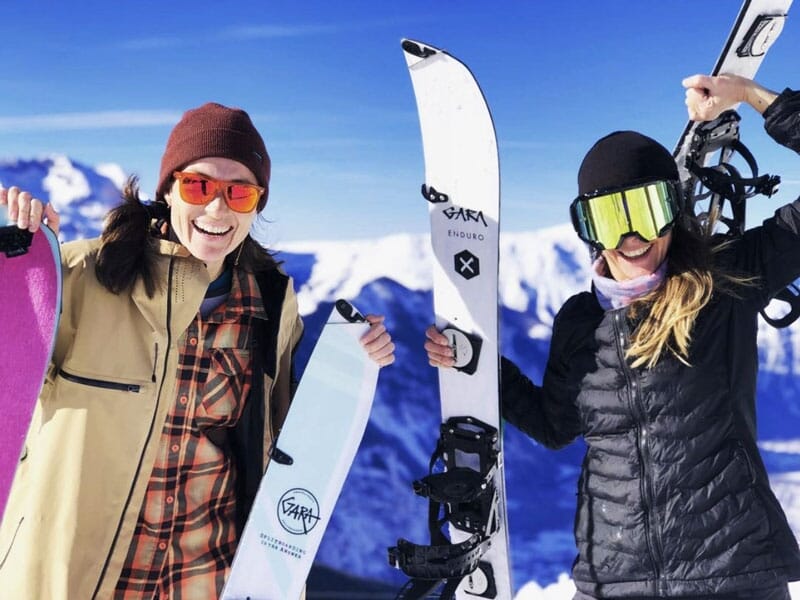 Deux snowboardeuses souriantes tenant leurs snowboards, prêtes à dévaler les pistes enneigées sous un ciel clair