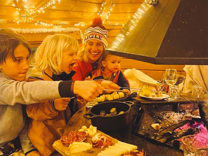 Une famille s'amuse autour d'un caquelon de fondue sur une table en bois dans une cabane chaleureusement éclairée, exprimant la joie et le partage