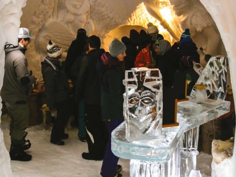 Des visiteurs en vêtements d'hiver explorant un bar de glace avec des sculptures et des meubles en glace éclairés, créant une atmosphère magique et givrée