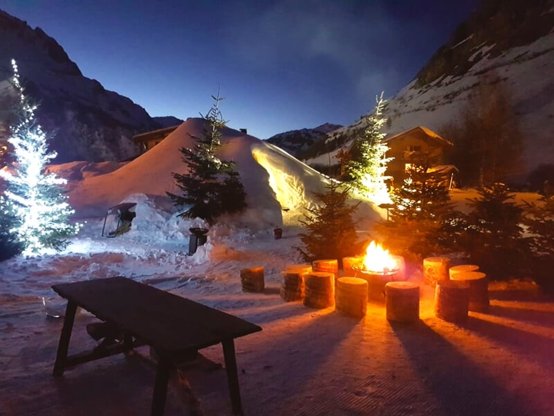 Un espace extérieur de détente la nuit en hiver, avec un feu de camp entouré de sièges en rondins et un éclairage festif près des arbres enneigés et des dômes