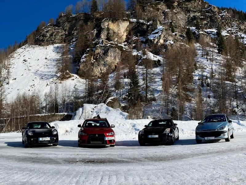 Quatre voitures de sport stationnées côte à côte sur une surface enneigée, avec une montagne rocheuse en arrière-plan sous un ciel bleu clair