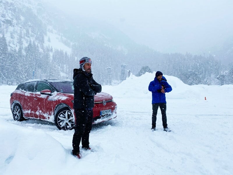 Deux hommes, l'un portant une veste rouge et l'autre une veste bleue, debout dans la neige à côté d'une voiture rouge couverte de neige pendant une chute de neige, avec des montagnes et des arbres en