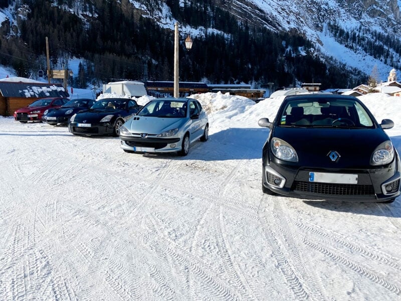 Plusieurs voitures stationnées sur une surface enneigée avec des traces de pneus visibles dans la neige, des bâtiments en bois et des montagnes en arrière-plan