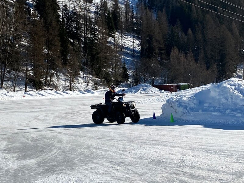 Un homme conduisant un quad noir sur une piste de glace avec des cônes de signalisation, entouré par un paysage hivernal avec des arbres et de la neige