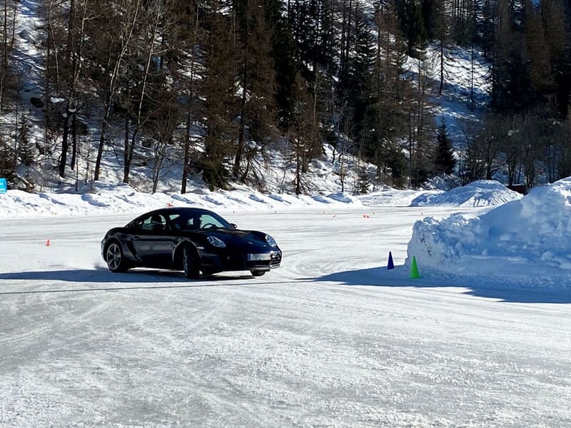 Une voiture de sport noire glissant sur une piste de glace avec des cônes de signalisation, entourée par un paysage hivernal enneigé et des arbres