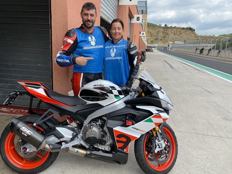 Deux pilotes souriants, l'un en combinaison de course rouge et l'autre en bleu, se tenant debout à côté d'une moto sportive Aprilia multicolore sur une piste de course