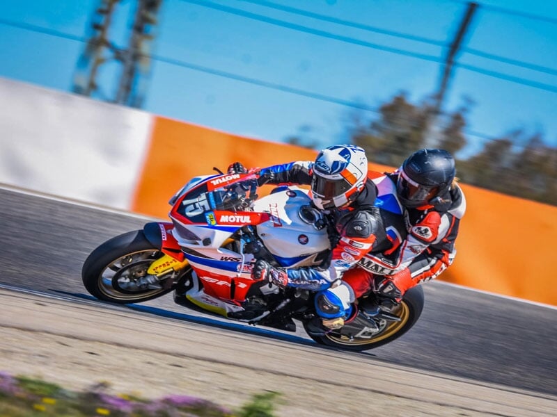 Pilote en combinaison de course rouge et blanche et son passager prenant un virage serré sur une moto de sport Honda lors d'une journée ensoleillée sur une piste de course
