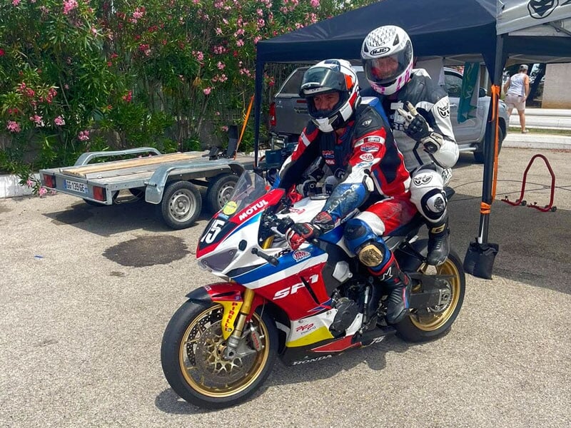 Deux pilotes en combinaison de course noire et blanche sur une moto de sport rouge et blanche Honda, garée devant une remorque et un abri avec des arbustes fleuris en arrière-plan