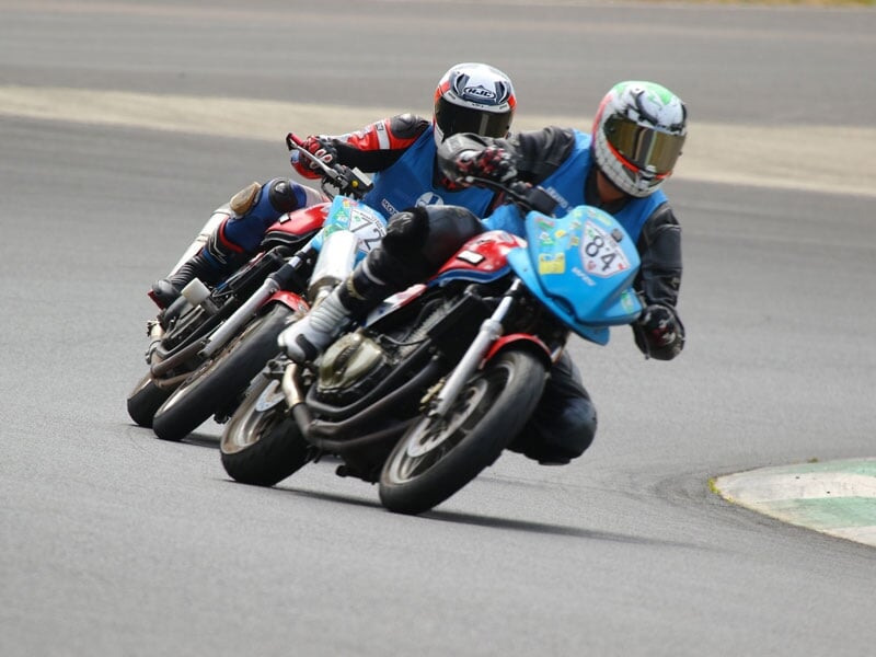 Deux coureurs de moto en tenue complète penchés dans un virage sur un circuit de course, entourés de verdure et de fleurs sauvages jaunes sous un ciel bleu clair