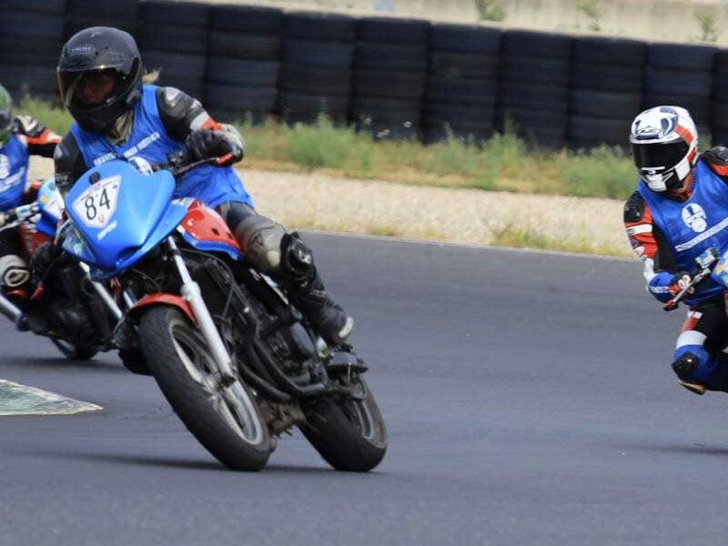 Des coureurs de moto sur une piste, avec un affichant de manière proéminente le numéro 84 en bleu, concentrés et penchés dans un virage à grande vitesse avec des barrières de pneus en arrière-plan