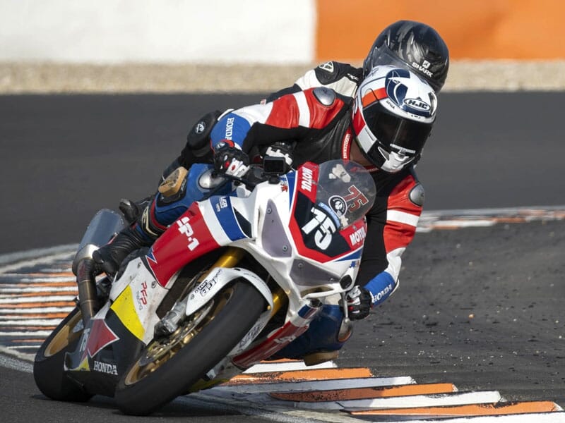 Pilote en combinaison rouge et bleue inclinant une moto sportive Honda dans un virage serré sur circuit