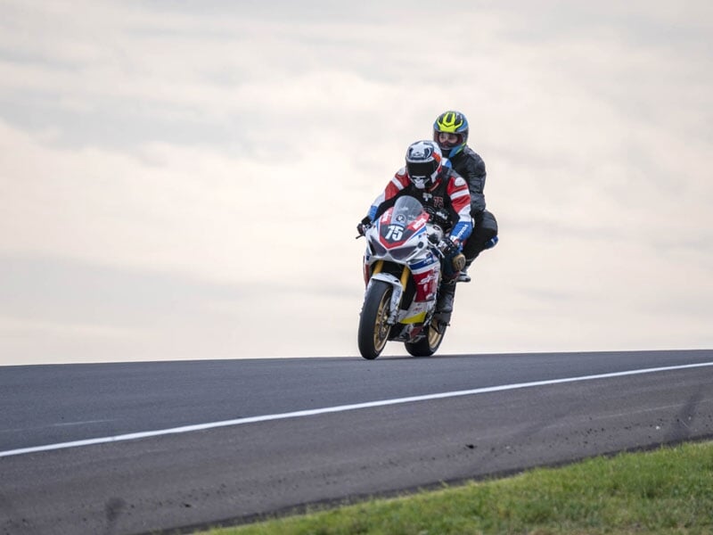 Moto sportive avec pilote et passager, roulant sur une piste, capturée au sommet d’une montée sous un ciel gris