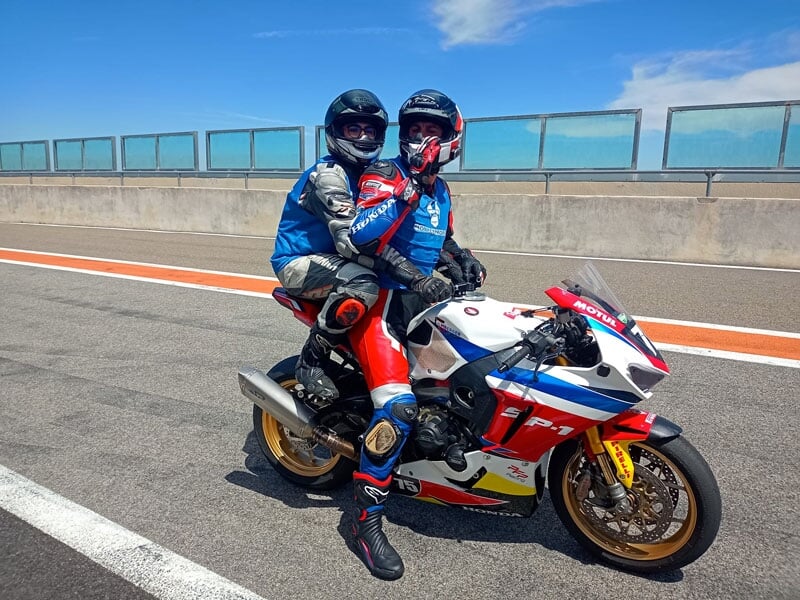 Deux motards en combinaison, assis sur une moto Honda sportive à l’arrêt sur une voie d’accès au circuit