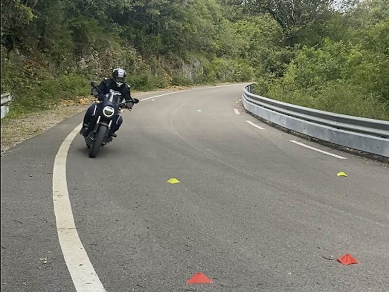 Motard en combinaison noire prenant un virage sur une route de montagne, entouré de cônes de signalisation colorés.
