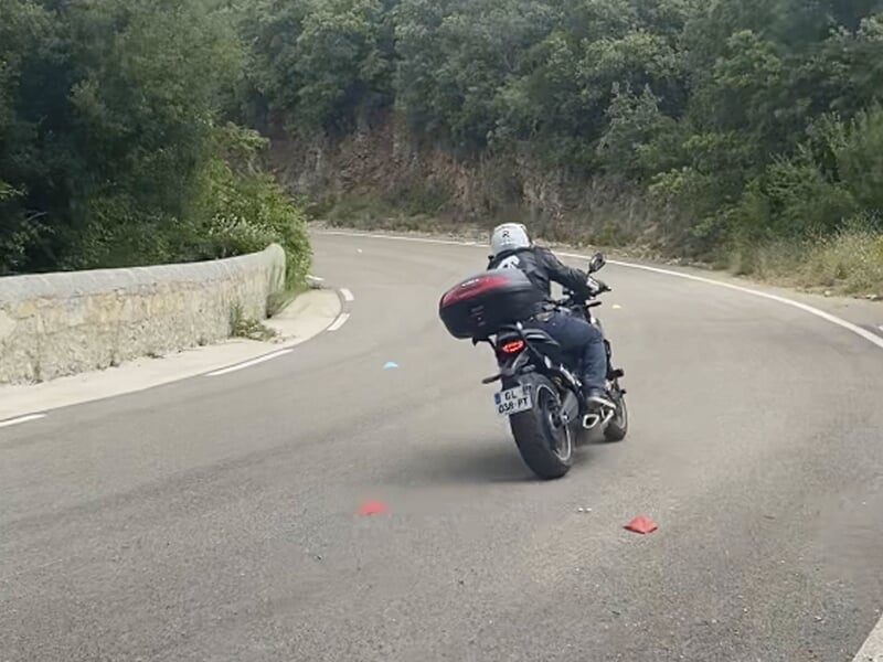 Vue arrière d'un motard avec un casque blanc, en train de négocier un virage sur une route sinueuse entourée de végétation.