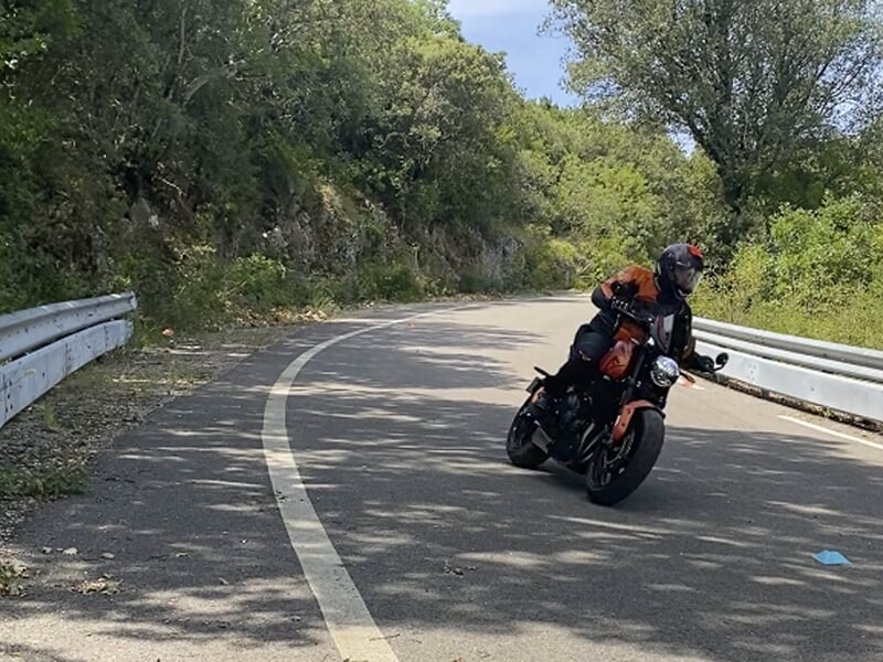 Motard en combinaison orange incliné dans un virage sur une route de montagne, encadré par des glissières de sécurité.
