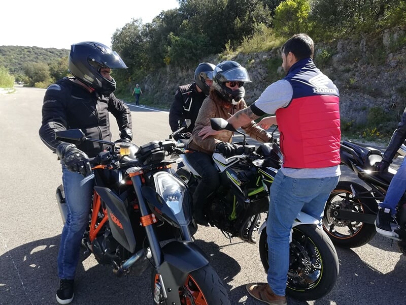 Groupe de motards à l'arrêt en train de discuter avec un instructeur en gilet rouge, avec leurs motos garées à proximité sur une route.
