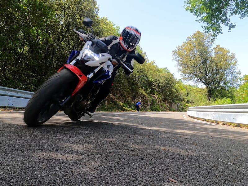 Motard en combinaison noire et casque rouge incliné de manière prononcée dans un virage serré sur une route de montagne.