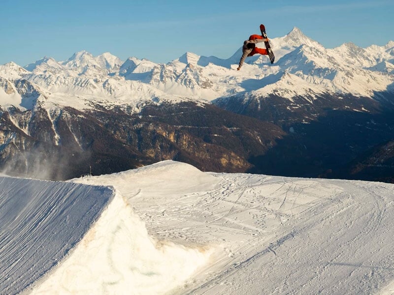 Snowboarder en plein saut avec vue panoramique sur une chaîne de montagnes enneigées, sous un ciel clair et ensoleillé