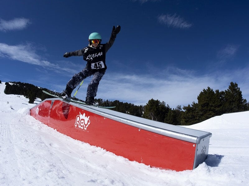 Enfant effectuant une figure sur une rampe de ski freestyle sous un ciel bleu éclatant. Les montagnes enneigées et les arbres bordent la piste