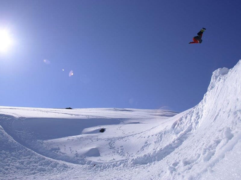 Snowboarder réalisant un saut spectaculaire dans les airs, laissant derrière lui une pente de neige poudreuse et un ciel dégagé