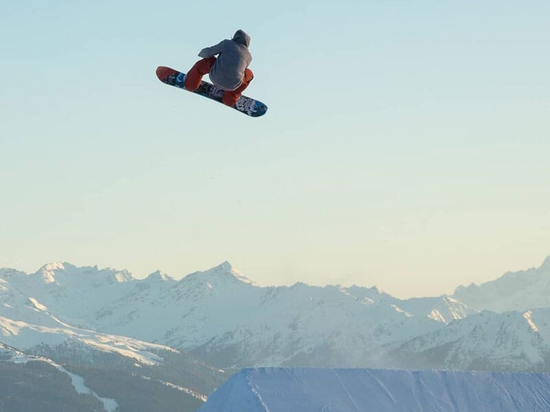 Snowboarder effectuant un saut acrobatique avec les montagnes enneigées en arrière-plan, le soleil couchant illuminant la scène