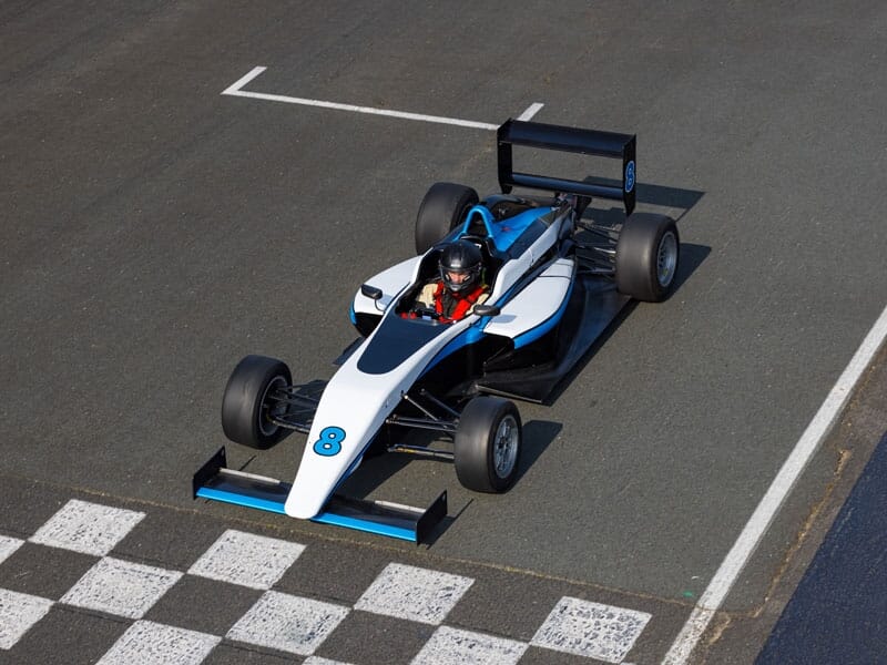 Voiture de course monoplace blanche et bleue franchissant la ligne d'arrivée, pilote en combinaison et casque noirs, vue aérienne