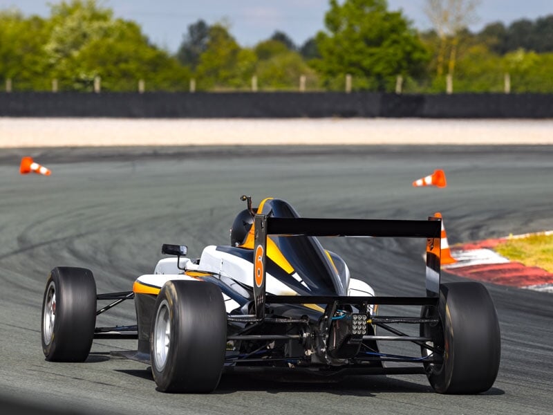 Monoplace de course sur circuit, en train de prendre un virage serré, pneus larges et carrosserie blanche avec détails noirs et oranges