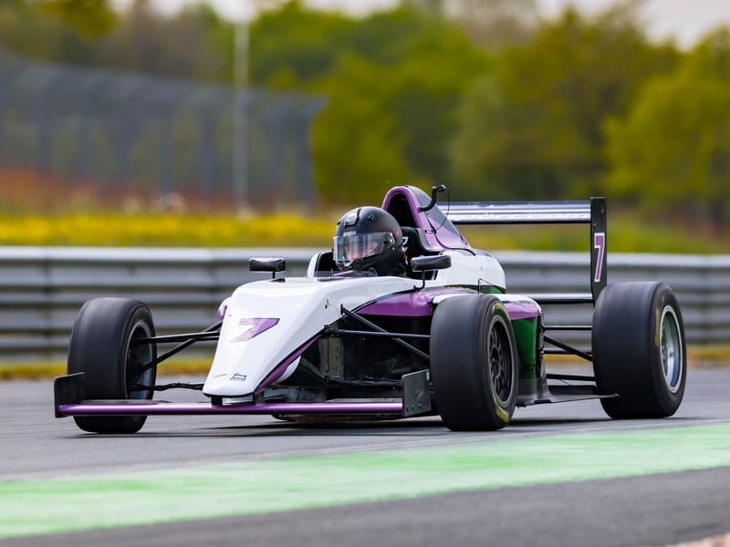 Voiture de course monoplace violette et blanche sur circuit, pilote en combinaison noire avec casque, entourée de verdure