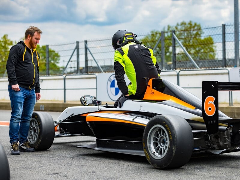 Pilote en combinaison et casque noir fluo montant dans une monoplace de course noire et orange, accompagnée par un mécanicien en veste noire